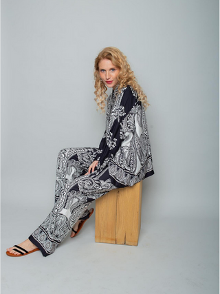 Frau sitzt auf Holzblock, trägt Emily Hose 201231 black/white mit Paisley-Muster, lockerem Schnitt und weitem Bein. Die Hose hat ein elastisches Bündchen und Seitentaschen, gefertigt aus weicher Viskose.