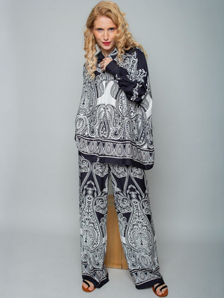 Person in Emily Bluse 8881-157910, eine oversized Hemdbluse mit XL-Saumweite und schmalem Ärmel. Schwarzer und weißer Paisley-Print, kombiniert mit weiten Hosen, offenen Sandalen und langem, welligem Haar.