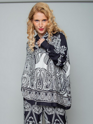 Person trägt die Emily Bluse 8881-157910 black/white, eine locker sitzende, langärmelige Tunika mit Paisleymuster, kombiniert mit passenden weiten Hosen. Sie hält den Kragen der Bluse sanft.