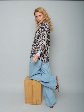 Emily Bluse 8812-154520 leo in lockerem Leopardenmuster mit leuchtenden Streifen an den Ärmelsäumen. Kombiniert mit weit geschnittenen Jeans und flachen Sandalen, knielang auf einem Holzblock posierend.