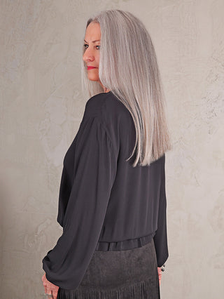 Frau in Profilansicht trägt die Emily Bluse 8900-158360 black, eine locker sitzende, langärmelige Bluse mit Bändern am Ausschnitt, kombiniert mit einem dunklen, fransigen Rock.