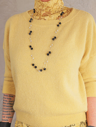 Person trägt die OFF#DLY Kette Riga black/silver, eine lange, mehrsträngige Halskette mit abwechselnden schwarzen und klaren facettierten Perlen, über einem senfgelben Strickpullover und gelbem Spitzenrollkragen.
