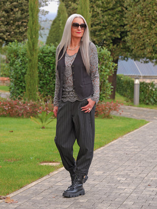 Ältere Frau trägt die OFF#DLY Weste Sirmione black: elegant fallend, leicht tailliert, mit Knopfleiste und dezenten Taschen, kombiniert über einer Spitzenbluse, auf einem Gartenweg spazierend.