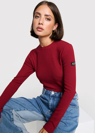ALIX Longsleeve Top bordeaux: Junge Frau trägt ein eng anliegendes, langärmeliges rotes Oberteil mit rundem Halsausschnitt und kleinem schwarzem Patch auf dem Ärmel, kombiniert mit zweifarbigen blauen Jeans.