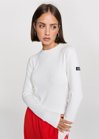 Junge Frau trägt das ALIX Longsleeve Top soft white, ein figurbetontes, weißes Rippstrick-Oberteil mit rundem Halsausschnitt und kleinem A|X Patch am linken Ärmel.