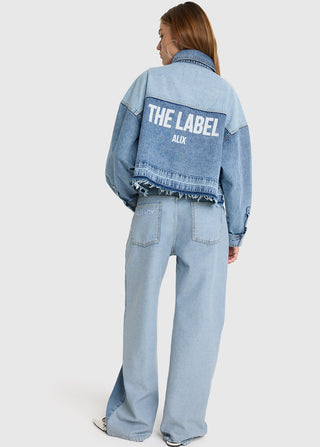 Rückenansicht eines Modells in der ALIX Denim Blouse light blue Denim. Die Bluse hat einen fransigen Saum und großen THE LABEL - ALIX Aufdruck, kombiniert mit passenden Jeans.
