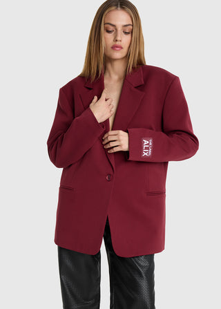 Modell trägt ALIX Oversized Blazer bordeaux mit Notch-Lapel und Logo-Patch am Ärmel, kombiniert mit schwarzen Hosen. Blazer hat eine einreihige Knopfleiste und zwei Fronttaschen.