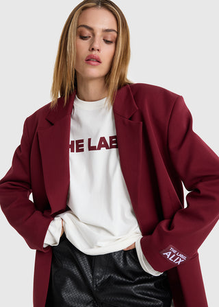 Junge Frau trägt ALIX Oversized Blazer bordeaux mit dezentem Label-Aufdruck am Ärmel. Kombiniert mit weißem T-Shirt und schwarzen, texturierten Kunstlederhosen. Hände in den Hosentaschen, neutraler Gesichtsausdruck.