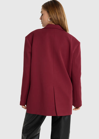 Person von hinten in einem ALIX Oversized Blazer bordeaux, der durch strukturierte Schulterpolster markant wirkt. Der Blazer fällt locker über schwarze, texturierte Hosen, mit entspannten Armen an den Seiten.