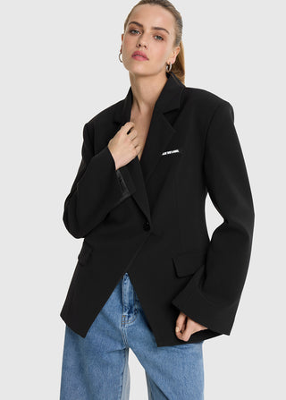 Person trägt den ALIX Fitted Blazer black, oversized mit Ein-Knopf-Front, leicht fallenden Schultern, breiten Ärmeln und dezentem ALIX THE LABEL Pin auf der Brust, kombiniert mit Jeans.