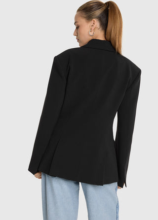 Person von hinten in einem ALIX Fitted Blazer black mit gepolsterten Schultern und ventiliertem Saum, kombiniert mit hellblauen Jeans, Haar im Pferdeschwanz, vor einem schlichten grauen Hintergrund.