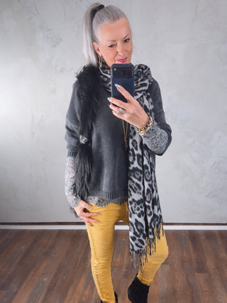 Frau mit hohem Pferdeschwanz trägt den OFF#DLY Leo Schal leo grau/schwarz über einem dunklen Pullover, kombiniert mit senfgelben Hosen und schwarzen Stiefeletten, in einem minimalistischen Raum.