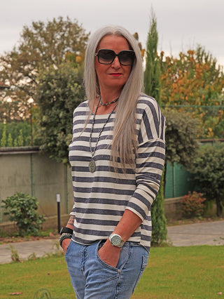 Frau im Garten trägt das OFF#DLY Ringel Glitzer Delia marine, ein gestreiftes Langarmshirt mit subtilem Glitzereffekt, kombiniert mit Jeans und Accessoires, die lässigen, eleganten Stil vermitteln.