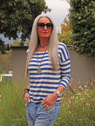 Die Frau trägt das OFF#DLY Ringel Glitzer Delia royalblue, ein blau-weiß gestreiftes Langarmshirt mit dezentem Glitzereffekt, kombiniert mit lässigen blauen Jeans und Accessoires im Garten.