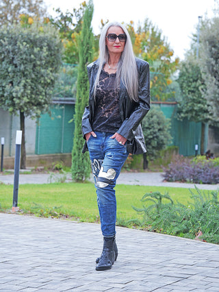 Frau trägt die OFF#DLY Destroy Denim Star midblue Jeans mit weißem Patch-Design, kombiniert mit schwarzem Lederblazer und Ankle Boots, in einem Garten oder Park stehend.