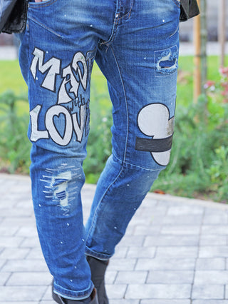 OFF#DLY Destroy Denin Star midblue Jeans mit Graffiti „MADE with LOVE“, Mickey-Patch, Rissen, und Farbspritzern, getragen mit dunklen Stiefeletten auf einem Weg im Freien.