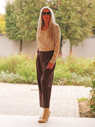 Ältere Frau in OFF#DLY Hose Moniga braun mit elegantem Schnitt und asymmetrischem Bund, posiert selbstbewusst in beige Spitzen-Top und tanfarbenen Stiefeletten, ergänzt durch goldene Accessoires.
