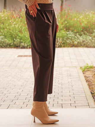 Die OFF#DLY Hose Moniga braun zeigt elegante, dunkelbraune, plissierte Vorderfalten, die am Knöchel verjüngen, gepaart mit beigefarbenen, spitzen Ankle Boots; Hand lässig in der Tasche.