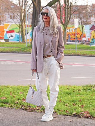 Frau in OFF#DLY White Denim Mira, entspannt geschnittene, leicht fließende weiße Denim-Hose, kombiniert mit Blazer und Sneakers, zeigt stilvolle Alltagstauglichkeit und modernen Charme.