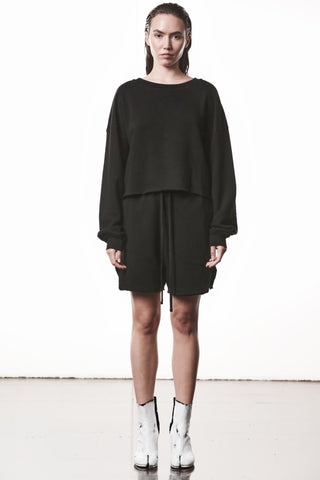 Junge Frau in einem locker geschnittenen, schwarzen Thom Krom WS496 Sweatshirt mit überschnittenen Schultern, leicht verkürztem Saum und geripptem Rundhalsausschnitt, kombiniert mit schwarzen Shorts und weißen Stiefeln.
