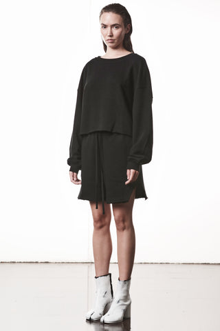 Frau trägt Thom Krom WST 432 Hose black: schwarze Mini-Rock-Shorts mit Kordelzug, seitlichen Schlitzen und lockerer Passform, kombiniert mit einem schwarzen Pullover und weißen Stiefeletten.