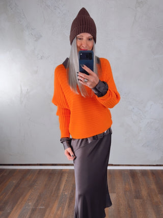 Person in einem hellen Raum mit Holzfußboden macht ein Spiegelselfie und trägt die OFF#DLY Mütze Anna schoko, kombiniert mit einem orangefarbenen Pullover, dunkler Bluse und langer, glänzender Rock.