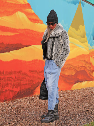 Person in OFF#DLY Denim Hella lightblue, lässige Jeans mit lockerer Passform, kombiniert mit schwarzen Boots und grauer Jacke, stehend vor einer farbenfrohen, abstrakten Wandmalerei.