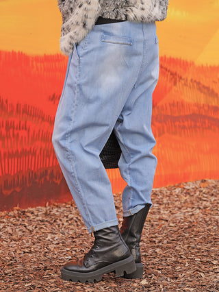 Person trägt OFF#DLY Denim Hella lightblue, locker sitzende, umgeschlagene Jeans, kombiniert mit schwarzen Stiefeln und einer schwarzen Tasche, stehend auf Holzspänen vor einer orange-roten Wand.