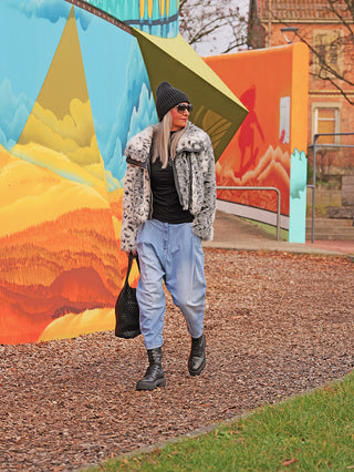 Eine Frau trägt die OFF#DLY Denim Hella lightblue, lässige Harem-Jeans mit weitem Schnitt und leicht zulaufendem Bein, kombiniert mit einem schwarzen Outfit und chunky Boots vor einem farbenfrohen Wandgemälde.