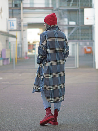 Person in einem Nuomeno Mantel Over Con mit modernem Karomuster in Blau und Grau, gehend in urbaner Umgebung. Der Mantel hat einen lässigen, oversized Schnitt, kombiniert mit hellen Jeans.
