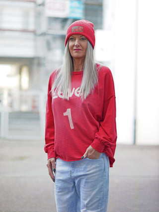 Person trägt OFF#DLY Sweat Level 1 red, ein locker geschnittenes rotes Sweatshirt mit einer großen weißen 1 und Teil des Wortes lover darauf, kombiniert mit hellen Jeans und roter Mütze.