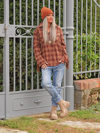 Person lehnt lässig am Metalltor, trägt die OFF#DLY Bluse Rodeo braun mit kariertem Muster und Fransen, kombiniert mit Jeans und Stiefeln in einem Gartenambiente.