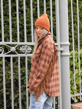 Person trägt OFF#DLY Bluse Rodeo braun: braun-karierte Flanellbluse mit langen Fransen an den Ärmeln, kombiniert mit orangefarbener Mütze und blauen Jeans, vermittelt lässigen, rustikalen Western-Stil.