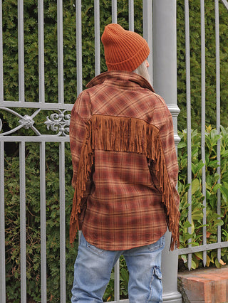 Person von hinten in rust-brauner OFF#DLY Bluse Rodeo mit Fransen, trägt blaue Jeans und orange Mütze, steht vor einem Metallzaun mit Pflanzen dahinter.