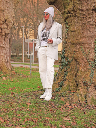 Person trägt OFF#DLY White Denim Nina, eine weiße Hose mit geradem Schnitt, in einem Park. Die moderne Silhouette ergänzt das minimalistische Outfit mit Blazer und Boots perfekt.