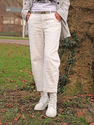 Person trägt OFF#DLY White Denim Nina: gerade, knöchellange, off-white Hose mit klassischen 5-Pockets, kombiniert mit Chunky Boots, Händen in den Taschen, vor Baumstamm und Laub am Boden.