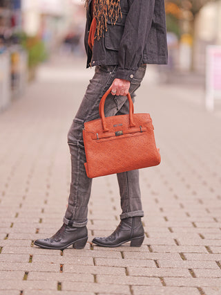 Die Sweet Years Logo Bag cognac wird von einer Person gehalten, die auf einem Gehweg steht. Die strukturierte, orangefarbene Handtasche hat geprägtes Muster und kurze Tragegriffe.