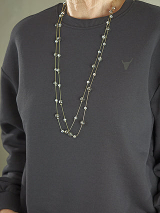 Die OFF#DLY Kette Riga grey/gold zeigt zwei lange, filigrane Ketten mit rauchgrauen und klaren Kristallperlen, getragen über einem dunklen Oberteil.