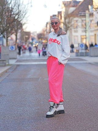 Person trägt den OFF#DLY Sweater Malibu white, ein off-shoulder Sweatshirt mit rotem Malibu California 1980 Schriftzug, kombiniert mit pinken Sweatpants und weißen Stiefeln.