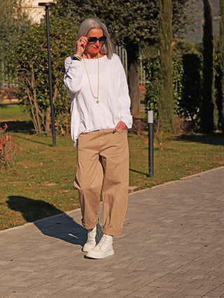 Eine Frau trägt den OFF#DLY Pulli Agathe, einen lockeren weißen Strickpullover mit feinem Strukturmuster und ausgefranstem Saum, kombiniert mit lässigen Accessoires auf einem Gartenweg.