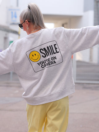 Person trägt den OFF#DLY Sweater Smile lightgrey mit großem gelbem Smiley und SMILE YOU’RE ON CAMERA-Schriftzug hinten, silberblonde Haare im Zopf, schwarze Sonnenbrille, gelbe Hose.