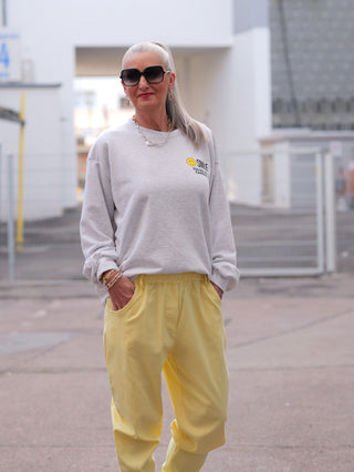 Ältere Person in OFF#DLY Sweater Smile lightgrey mit Smiley-Print, lässigem Schnitt und silbernen Accessoires, in entspannter Pose draußen, verkörpert modernen, zeitlosen Street-Style.