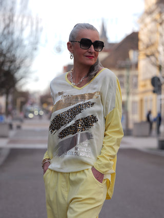 Silberhaarige Frau trägt das OFF#DLY LS Nora gelb, ein lässiges Longsleeve mit V-Ausschnitt, metallicen Elementen und inspirierendem Schriftzug, kombiniert mit passender gelber Hose auf ruhiger Straße.