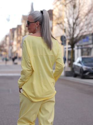 Eine Person trägt das OFF#DLY LS Nora gelb, ein locker geschnittenes Longsleeve mit V-Ausschnitt, während sie in einer Stadtstraße steht. Sichtbar sind auch ihre hochgebundene silberne Haarpracht und Sonnenbrille.