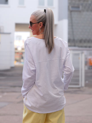 Person von hinten in lockerem OFF#DLY LS Nora white Longsleeve, kombiniert mit modischen Details wie V-Ausschnitt und stilvollem Frontprint, steht in urbaner Umgebung.