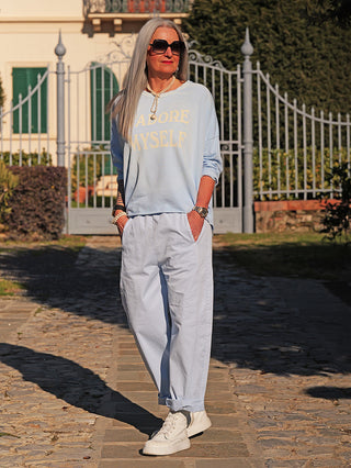 Mature Frau trägt das OFF#DLY Sweat J’adore lightblue mit dem Slogan „ADORE MYSELF“. Kombiniert es mit lässigen, passenden Hosen, weißen Sneakers, Perlenkette und Sonnenbrille beim Spaziergang.