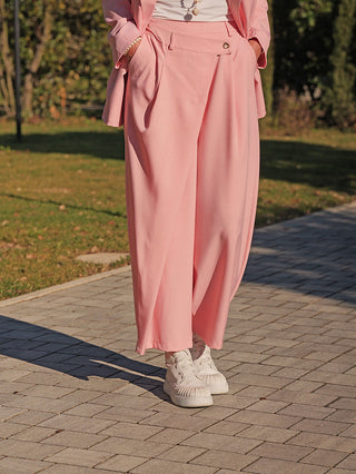 Person trägt OFF#DLY Hose Lisa rose, eine locker fallende, roséfarbene Hose mit seitlichen Taschen und weitem Bein, kombiniert mit weißen Sneakers auf einem gepflasterten Weg im Freien.