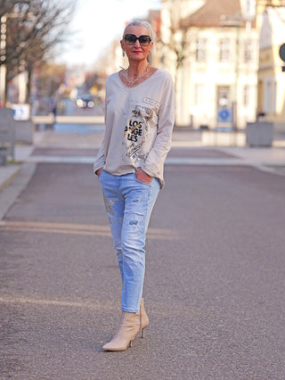 Eine Frau trägt die OFF#DLY Destoy Denim Queen lightblue Jeans mit Used-Effekten und glitzernden Applikationen, kombiniert mit einem beigen LOS ANGELES Shirt und passenden Stiefeletten.