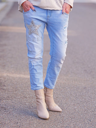 OFF#DLY Destroy Denim Queen lightblue Jeans mit funkelndem Stern und „QUEEN“-Schriftzug, leichte Used-Effekte. Kombiniert mit spitzen, nude-farbenen Stiefeletten. Hände lässig in den Taschen.