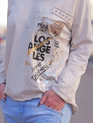 Nahaufnahme des OFF#DLY LS Mia beige Langarmshirts mit LOS ANGELES und FOREVER DREAMER Schriftzug, Metallic-Folien, Pailletten und Nieten. Hände in Taschen einer hellblauen Jeans.